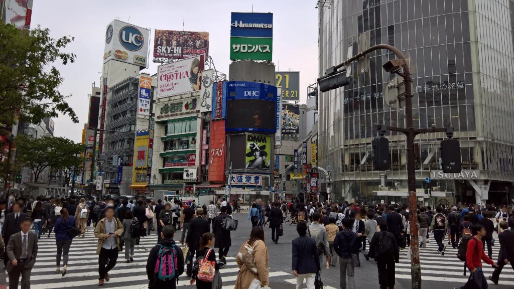 Picture of Shibuya Crossing. A lot of people walking across the street. High buildings with commercials.