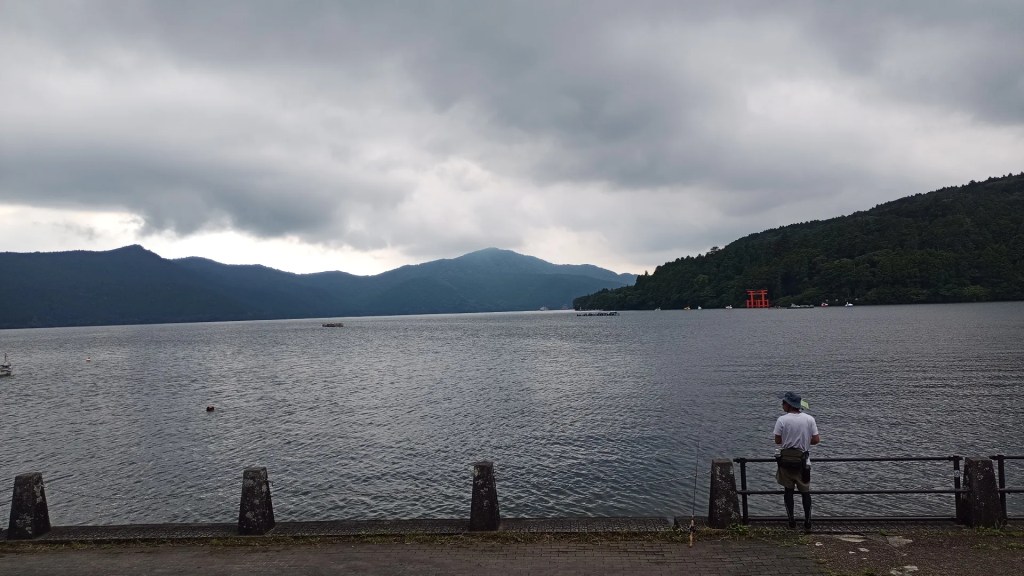 A view of Lake Ashinoko, Hakone. The water is quiet. A fisherman standing at the shore.