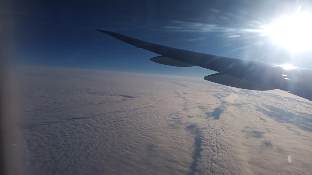 A view above the clouds out of an airplane window with the sun in front.